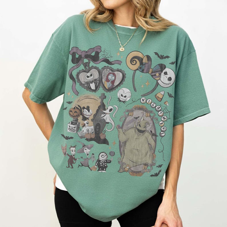 May include: A sage green short-sleeve t-shirt featuring a graphic print of characters from The Nightmare Before Christmas, including Jack Skellington, Sally, and Oogie Boogie, alongside various Halloween-themed icons like bats and a haunted house.