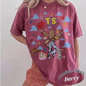 May include: A berry-colored short-sleeve t-shirt featuring a graphic print of a person playing a guitar, surrounded by blue clouds, stars, flowers, a red hat, a brown cowboy hat, and blocks with the text TS. The shirt has a crew neckline.