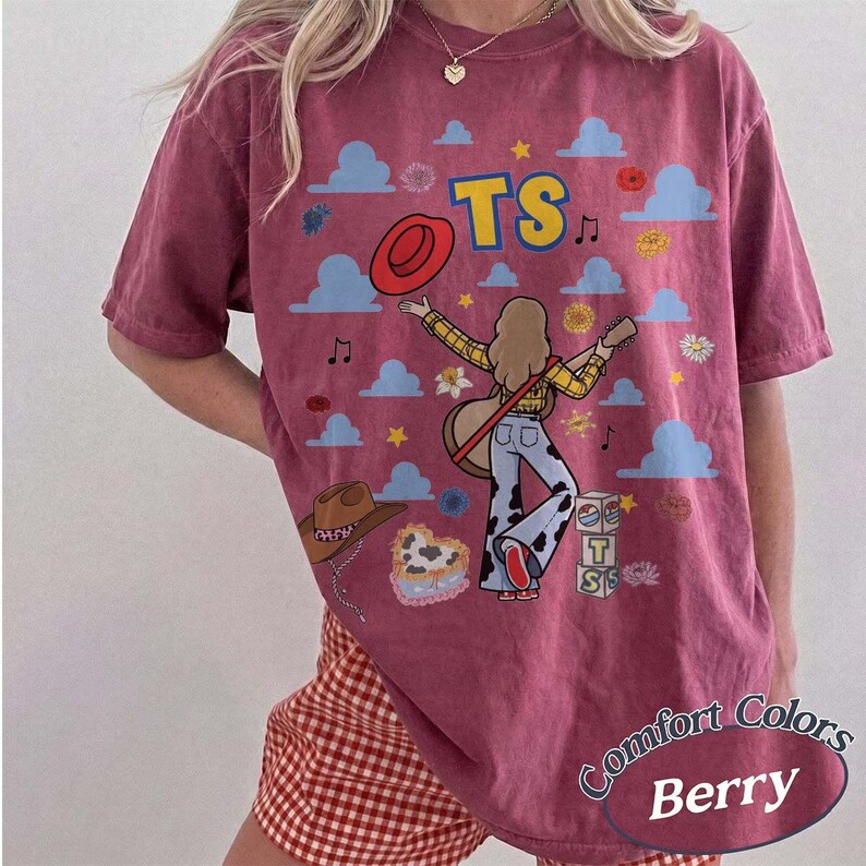 May include: A berry-colored short-sleeve t-shirt featuring a graphic print of a person playing a guitar, surrounded by blue clouds, stars, flowers, a red hat, a brown cowboy hat, and blocks with the text TS. The shirt has a crew neckline.