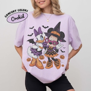 Może przedstawiać: Jasnofioletowy t-shirt z grafiką przedstawiającą Daisy Duck i Minnie Mouse w kostiumach na Halloween. Daisy ma na sobie skrzydła nietoperza i fioletową sukienkę, a Minnie nosi kapelusz czarownicy i okulary. Postacie otaczają małe dynie i nietoperze.