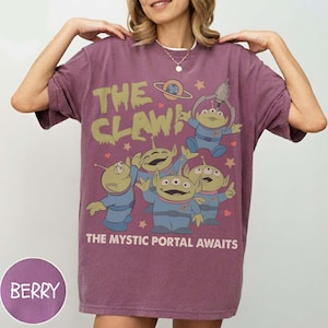 May include: Berry-colored t-shirt featuring a graphic of the Little Green Men from Toy Story. The design includes the text "THE CLAW" and "THE MYSTIC PORTAL AWAITS". The aliens are depicted in blue uniforms with a space-themed background.