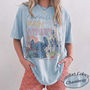 May include: Light blue t-shirt with a graphic featuring Stitch, a castle, and the text "Meet Me On Main Street." The shirt has a relaxed fit and short sleeves. The bottom right corner has the text "Comfort Colors Chambray."