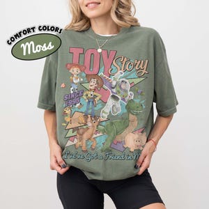 May include: A moss green short-sleeve t-shirt featuring a graphic print of Toy Story characters including Woody, Buzz Lightyear, Jessie, Rex, Slinky Dog, and Hamm. The text reads "TOY Story", "SINCE 1995", and "I've ve Got a Friend in Me".