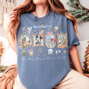May include: A blue t-shirt featuring various Disney characters in framed illustrations. The shirt includes the text "Walt Disney World" and "2026", along with the phrase "The Most Magical Place on Earth".