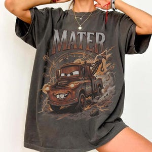 Puede incluir: Camiseta gris oscuro con una imagen de dibujos animados de Mater de la pel&iacute;cula Cars. La palabra "MATER" est&aacute; impresa encima de la imagen en un estilo desgastado. El dise&ntilde;o incluye rayos y un fondo circular.