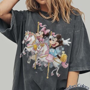 Op de afbeelding: Donkergrijs T-shirt met een carrouselontwerp met Mickey en Minnie Mouse op witte paarden. De afbeelding bevat roze en blauwe details. Het shirt heeft een vintage, vervaagde uitstraling en de tekst "Comfort Colors Pepper" staat rechtsonder.