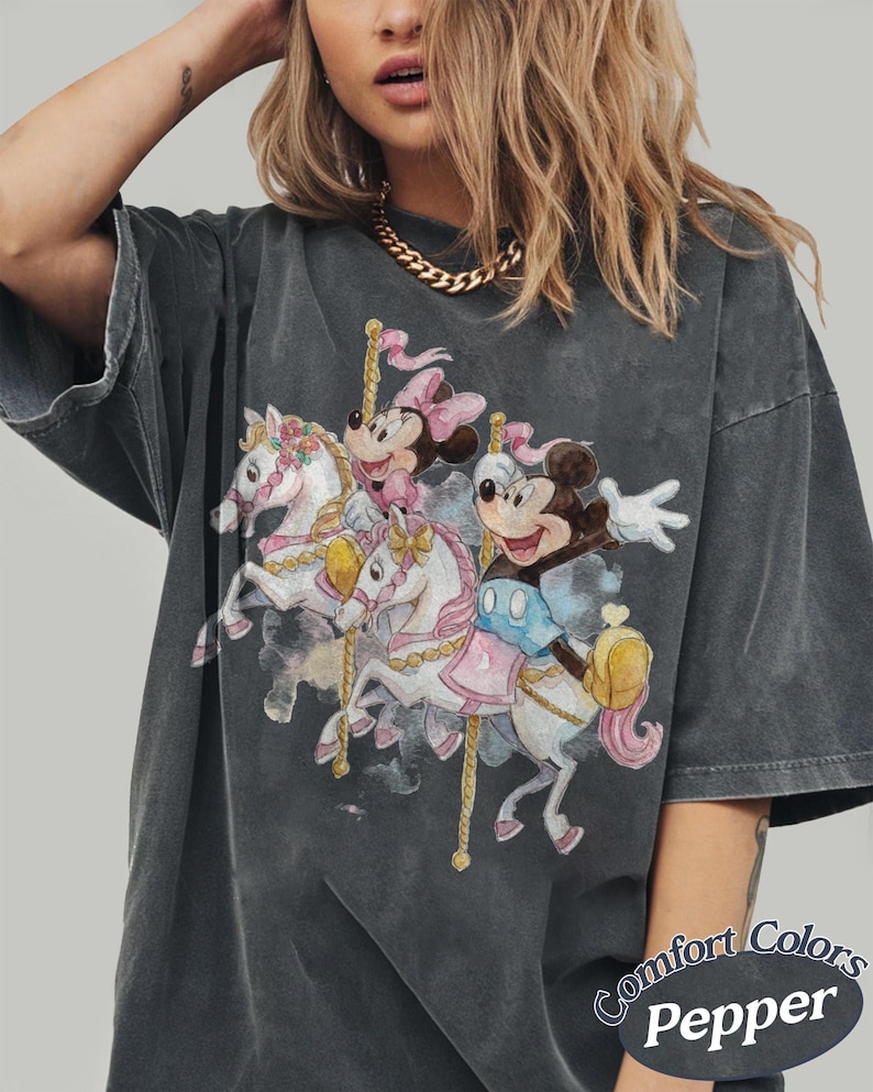 Op de afbeelding: Donkergrijs T-shirt met een carrouselontwerp met Mickey en Minnie Mouse op witte paarden. De afbeelding bevat roze en blauwe details. Het shirt heeft een vintage, vervaagde uitstraling en de tekst "Comfort Colors Pepper" staat rechtsonder.