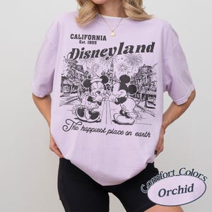 Puede incluir: Una camiseta de color lila claro con un gr&aacute;fico de Disneyland. El gr&aacute;fico presenta a Mickey y Minnie Mouse, fuegos artificiales y el texto "CALIFORNIA Est. 1955 Disneyland The happiest place on earth". La camiseta est&aacute; etiquetada como "Comfort Colors Orchid".