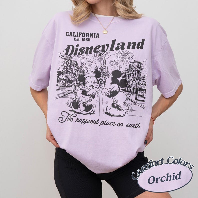 Puede incluir: Una camiseta de color lila claro con un gr&aacute;fico de Disneyland. El gr&aacute;fico presenta a Mickey y Minnie Mouse, fuegos artificiales y el texto "CALIFORNIA Est. 1955 Disneyland The happiest place on earth". La camiseta est&aacute; etiquetada como "Comfort Colors Orchid".