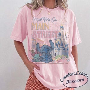 May include: A light pink t-shirt with the text "Meet Me On Main Street" and a graphic of Stitch and a castle. The shirt is from Comfort Colors and is in the Blossom color.