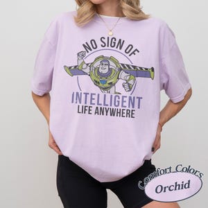 May include: A light purple t-shirt with a graphic of Buzz Lightyear and the text "NO SIGN OF INTELLIGENT LIFE ANYWHERE." The shirt is a Comfort Colors Orchid. The design features Buzz Lightyear in his space suit with his wings extended.