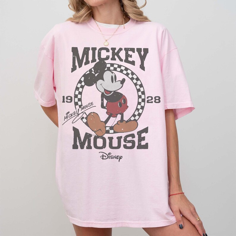 May include: A light pink t-shirt featuring a distressed graphic of Mickey Mouse. The design includes the words "Mickey Mouse" in a bold font, along with the years "19" and "28". The Disney logo is at the bottom.