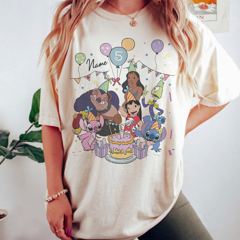 Pu&ograve; includere: T-shirt color crema con una grafica colorata di personaggi di cartoni animati che festeggiano un compleanno. Il design include palloncini, una torta e il numero 5. La parola "Name" &egrave; visibile.