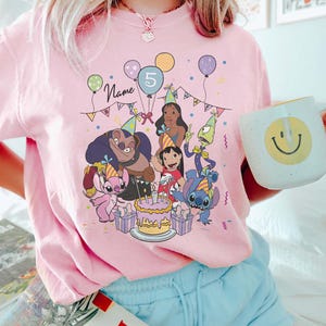 Pu&ograve; includere: T-shirt rosa chiaro con una grafica colorata di personaggi dei cartoni animati che festeggiano un compleanno. Il design include palloncini, una torta e il numero 5. &Egrave; visibile anche la parola "Name".