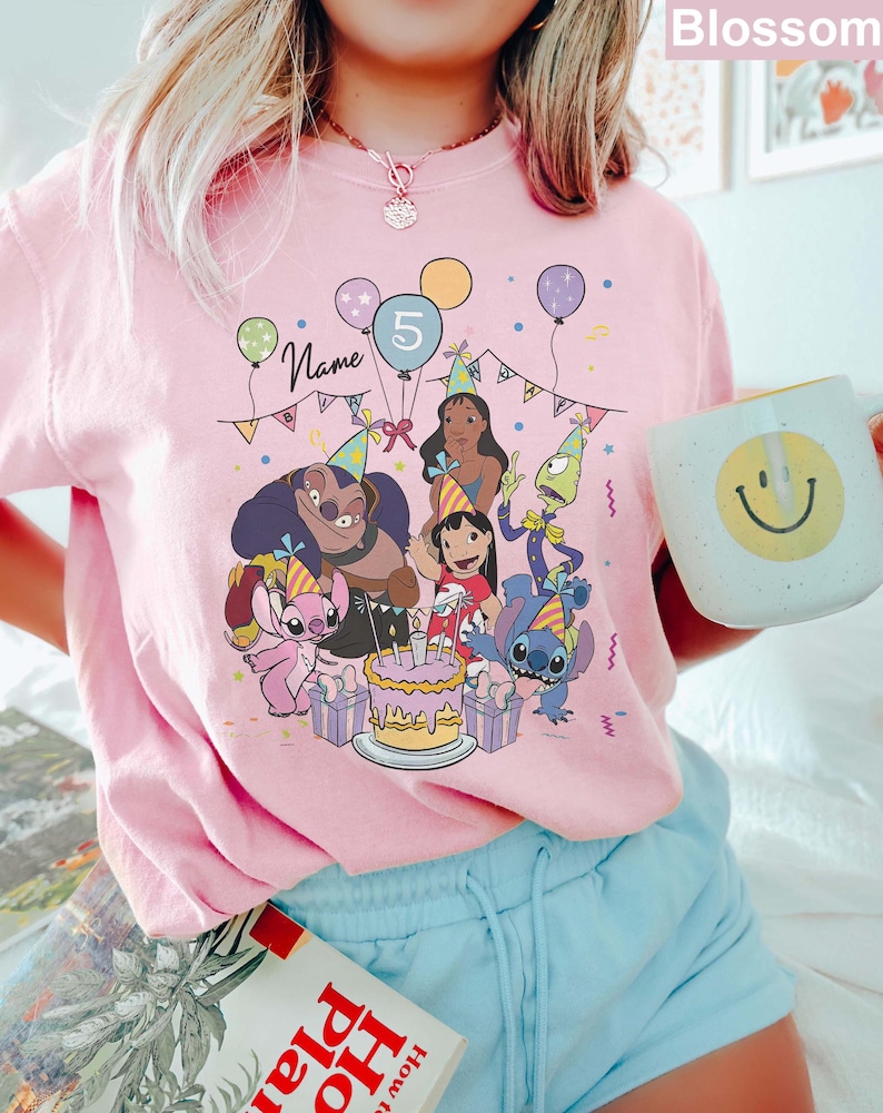 Pu&ograve; includere: T-shirt rosa chiaro con una grafica colorata di personaggi dei cartoni animati che festeggiano un compleanno. Il design include palloncini, una torta e il numero 5. &Egrave; visibile anche la parola "Name".