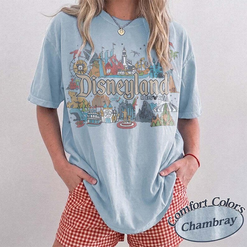 May include: Light blue t-shirt with a Disneyland Resort graphic. The graphic features various park attractions and the Disneyland Resort logo. The shirt is a Comfort Colors Chambray style.