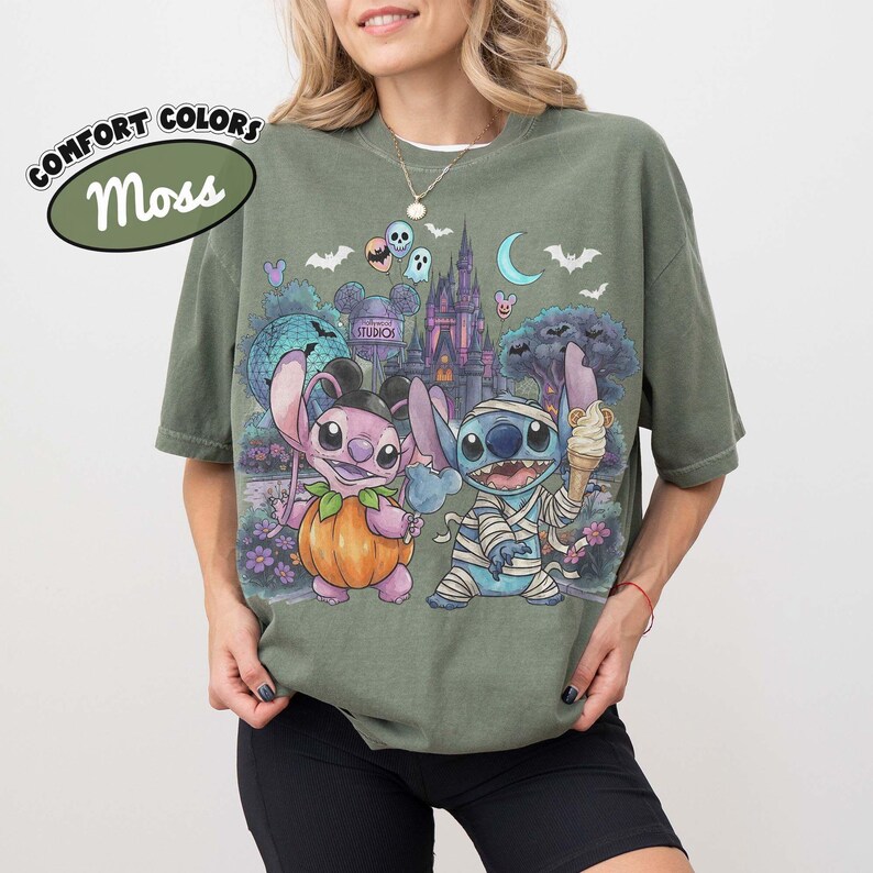 May include: A moss green t-shirt featuring a graphic print of two blue alien characters in Halloween costumes. One character is dressed as a pumpkin, the other as a mummy holding an ice cream cone. The background shows a castle, bats, and moons.