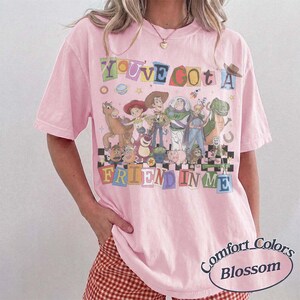 May include: A light pink short-sleeve t-shirt featuring a graphic print of various Toy Story characters. The text reads YOU'VE GOT A FRIEND IN ME in colorful, block-style letters. The design includes Woody, Buzz Lightyear, and other figures.