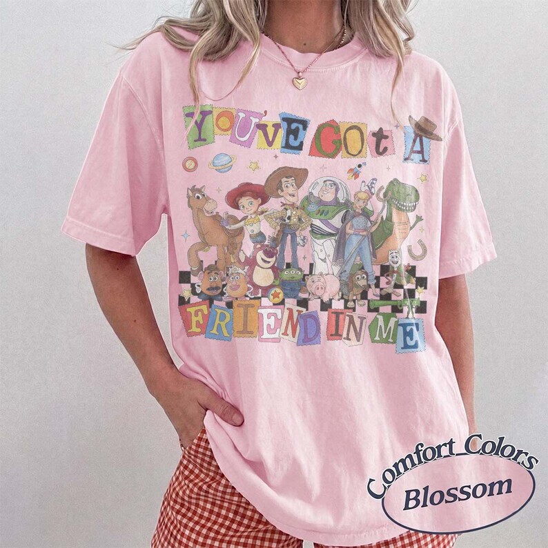 May include: A light pink short-sleeve t-shirt featuring a graphic print of various Toy Story characters. The text reads YOU'VE GOT A FRIEND IN ME in colorful, block-style letters. The design includes Woody, Buzz Lightyear, and other figures.