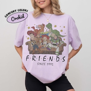 May include: An orchid-colored t-shirt featuring a graphic print of characters from Toy Story sitting on a couch. The text reads "F R I E N D S" and "SINCE 1995" below the illustration. A small oval label in the top left corner says "COMFORT COLORS Orchid".