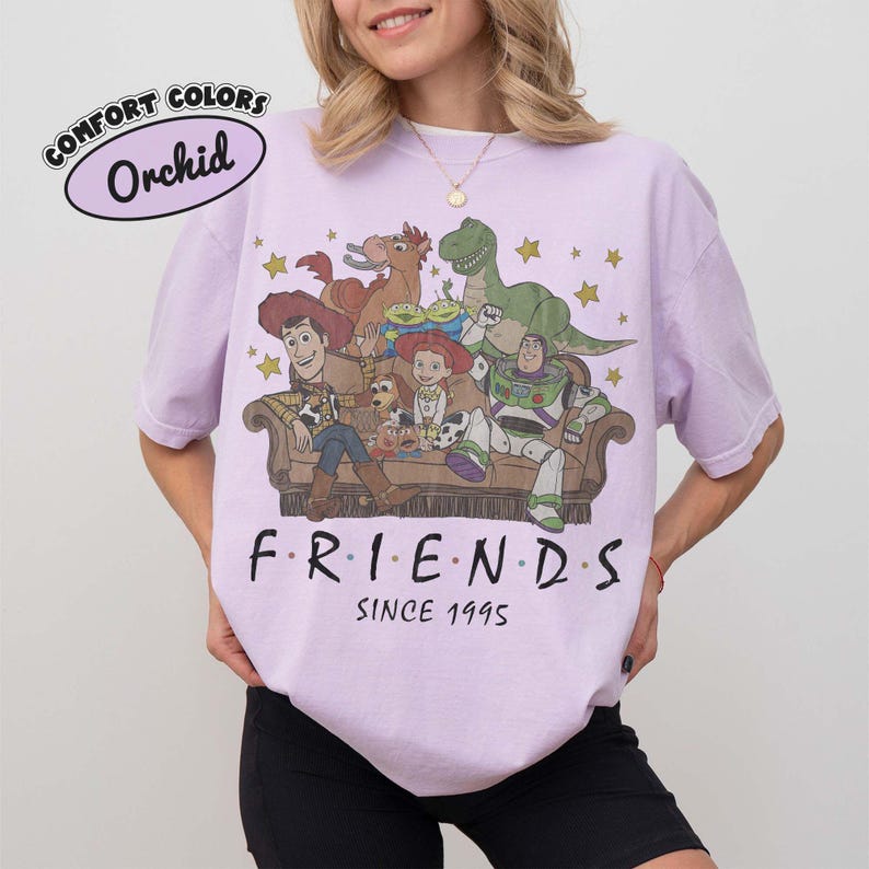May include: An orchid-colored t-shirt featuring a graphic print of characters from Toy Story sitting on a couch. The text reads "F R I E N D S" and "SINCE 1995" below the illustration. A small oval label in the top left corner says "COMFORT COLORS Orchid".