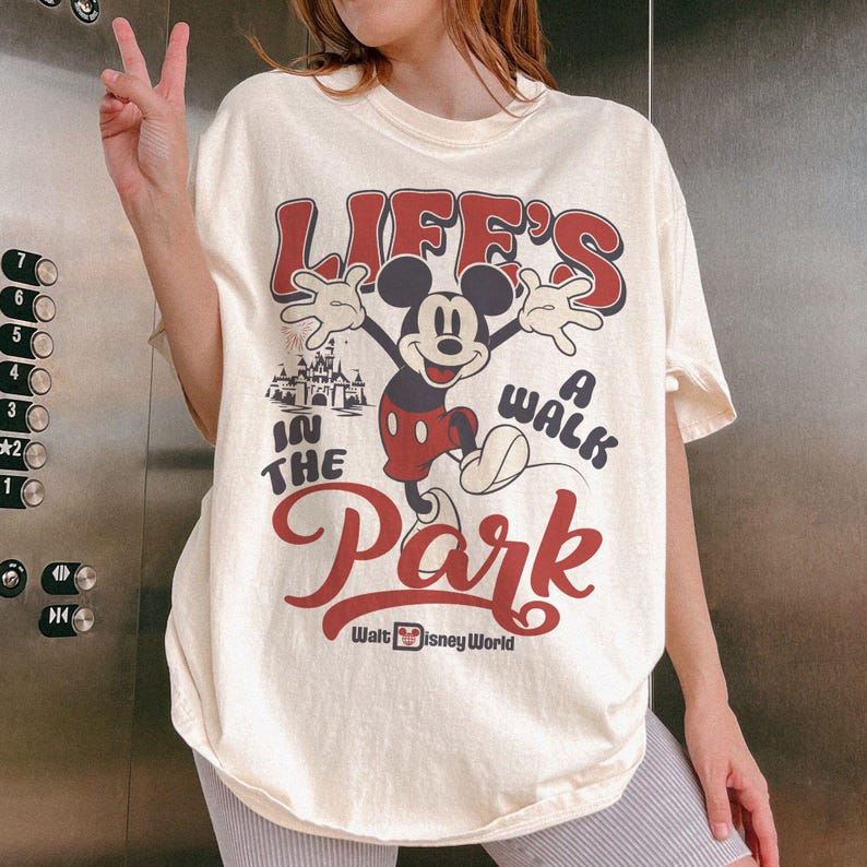 May include: Cream-colored t-shirt featuring Mickey Mouse with the text "LIFE'S A WALK IN THE PARK" in red and black. The design includes a castle illustration and the words "Walt Disney World".