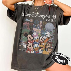 May include: A charcoal gray short-sleeve t-shirt featuring a graphic of Walt Disney World Est. 1971 with Mickey Mouse, Minnie Mouse, Donald Duck, Daisy Duck, Goofy, Pluto, Chip, and Dale taking a selfie in front of the park landmarks.