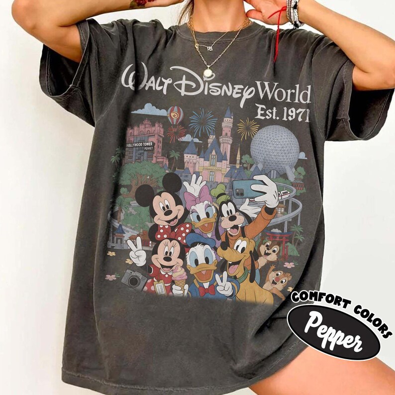 May include: A charcoal gray short-sleeve t-shirt featuring a graphic of Walt Disney World Est. 1971 with Mickey Mouse, Minnie Mouse, Donald Duck, Daisy Duck, Goofy, Pluto, Chip, and Dale taking a selfie in front of the park landmarks.
