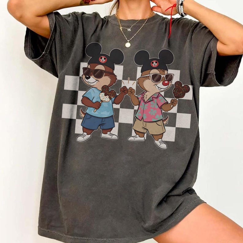 May include: Dark gray t-shirt featuring a cartoon illustration of two chipmunks wearing sunglasses and Mickey Mouse hats. One chipmunk is wearing a blue shirt and the other a pink floral shirt, both holding ice cream treats. A black and white checkered pattern is in the background.