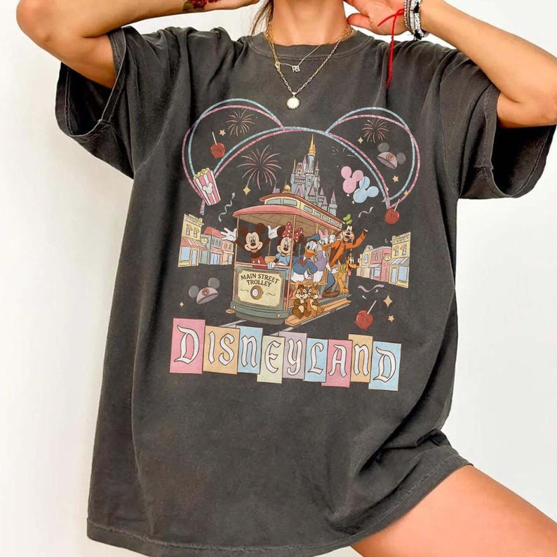 May include: Dark gray t-shirt featuring a Disneyland design. The graphic includes Mickey and Minnie Mouse, Donald Duck, and Goofy riding a trolley. The word "Disneyland" is displayed in colorful letters below the trolley.