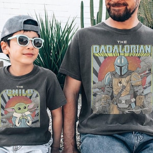 Puede incluir: Dos camisetas grises antracita a juego. Una camiseta presenta "THE CHILD" con un gr&aacute;fico de Grogu, la otra dice "THE DADALORIAN" con un gr&aacute;fico mandaloriano. Ambas camisetas tienen un aspecto vintage y desgastado.