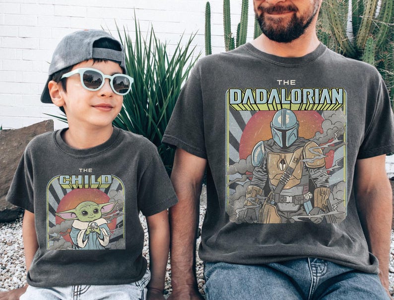 Puede incluir: Dos camisetas grises antracita a juego. Una camiseta presenta "THE CHILD" con un gr&aacute;fico de Grogu, la otra dice "THE DADALORIAN" con un gr&aacute;fico mandaloriano. Ambas camisetas tienen un aspecto vintage y desgastado.