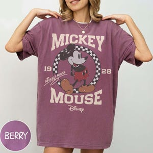 May include: Berry-colored t-shirt featuring a vintage Mickey Mouse graphic. The design includes the words "Mickey Mouse" and "Disney", along with the years "19" and "28". The graphic is set within a checkered circle.