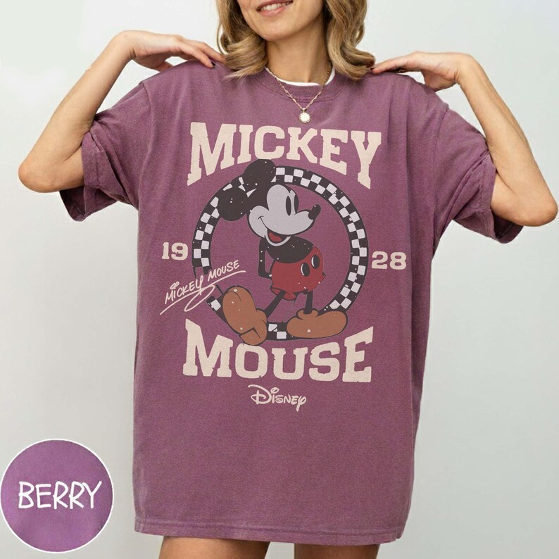 May include: Berry-colored t-shirt featuring a vintage Mickey Mouse graphic. The design includes the words "Mickey Mouse" and "Disney", along with the years "19" and "28". The graphic is set within a checkered circle.
