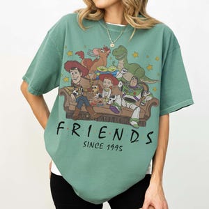 May include: A sage green short-sleeve t-shirt featuring a graphic print of Toy Story characters sitting on a couch. The text "FRIENDS" and "SINCE 1995" is printed in black below the illustration. The design includes Woody, Buzz Lightyear, Jessie, Rex, Bullseye, Slinky Dog, Mr. Potato Head, and the green aliens.