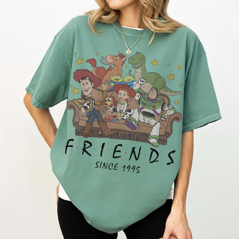May include: A sage green short-sleeve t-shirt featuring a graphic print of Toy Story characters sitting on a couch. The text "FRIENDS" and "SINCE 1995" is printed in black below the illustration. The design includes Woody, Buzz Lightyear, Jessie, Rex, Bullseye, Slinky Dog, Mr. Potato Head, and the green aliens.