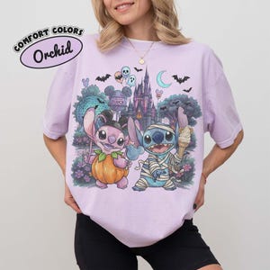 May include: A light purple t-shirt featuring a graphic print of two blue alien characters in Halloween costumes. One character is dressed as a pumpkin, the other as a mummy holding an ice cream cone. The background shows a castle, bats, and balloons.