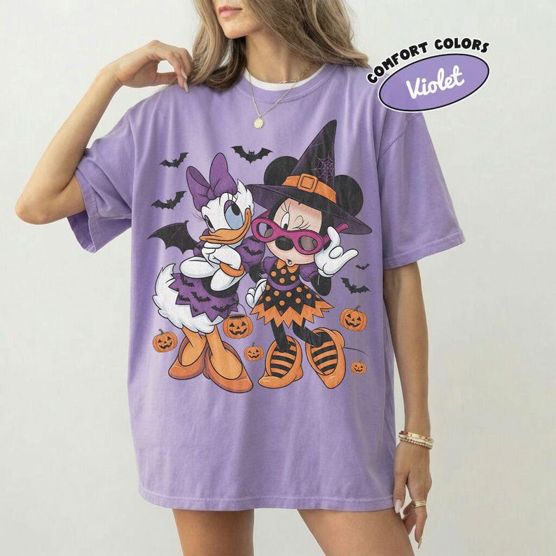 Może przedstawiać: Jasnofioletowy t-shirt z kr&oacute;tkim rękawem z grafiką przedstawiającą Daisy Duck i Minnie Mouse w kostiumach na Halloween. Wz&oacute;r zawiera nietoperze, pomarańczowe dynie oraz napis Comfort Colors Violet. Koszulka ma okrągły dekolt.