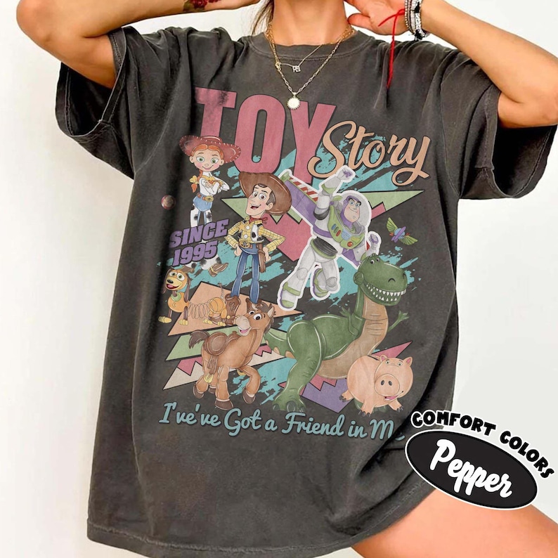 May include: A charcoal gray short-sleeve t-shirt featuring a graphic print of Toy Story characters including Woody, Buzz Lightyear, Jessie, Rex, Slinky Dog, and Hamm. The text reads Toy Story, Since 1995, and I've got a Friend in Me.