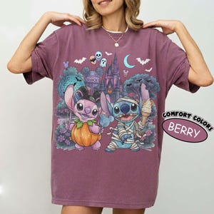 May include: A berry-colored t-shirt featuring a graphic print of two cartoon characters in Halloween costumes. One character is dressed as a pumpkin, and the other is wrapped in bandages like a mummy holding an ice cream cone. The background shows a castle, bats, and a crescent moon. The text COMFORT COLORS BERRY is visible.