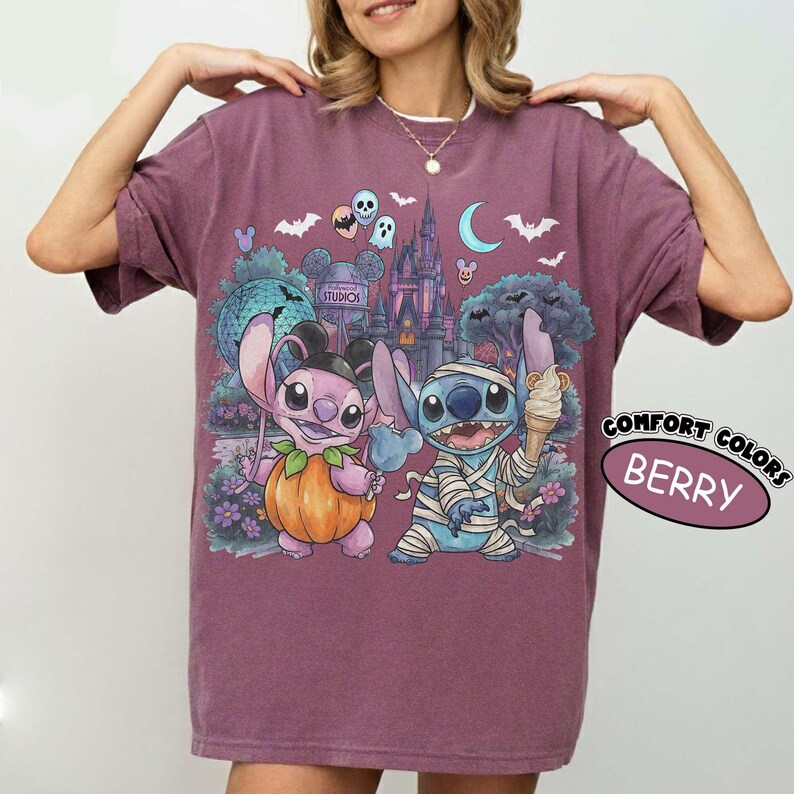 May include: A berry-colored t-shirt featuring a graphic print of two cartoon characters in Halloween costumes. One character is dressed as a pumpkin, and the other is wrapped in bandages like a mummy holding an ice cream cone. The background shows a castle, bats, and a crescent moon. The text COMFORT COLORS BERRY is visible.