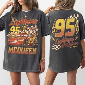 Może przedstawiać: Ciemnoszara koszulka Lightning McQueen z grafiką postaci i numerem 95. Projekt z przodu zawiera tekst "Lightning McQueen" i "Cars". Tył koszulki r&oacute;wnież wyświetla numer 95 i imię postaci.