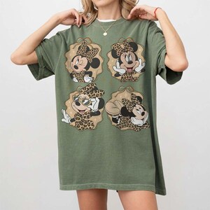 May include: Olive green t-shirt featuring four Minnie Mouse designs with leopard print bows and outfits. Each Minnie Mouse is set within a tan-colored, scalloped frame. The shirt has short sleeves and a relaxed fit.