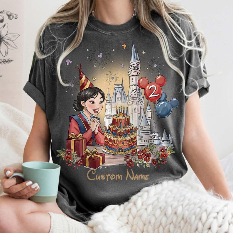 May include: Dark grey t-shirt with a birthday-themed cartoon illustration. The design includes a cake, gifts, a castle, balloons, and the text "Custom Name". The cartoon woman wears a party hat. A fun, celebratory design.