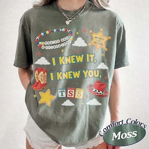 May include: A moss green short-sleeve t-shirt featuring a graphic print with the text I KNEW IT, I KNEW YOU, TS5, INFINITY AND BEYOND, and YOU'VE GOT A FRIEND IN ME. The design includes a beaded bracelet, a gold star, red cowboy boots, and a red hat.