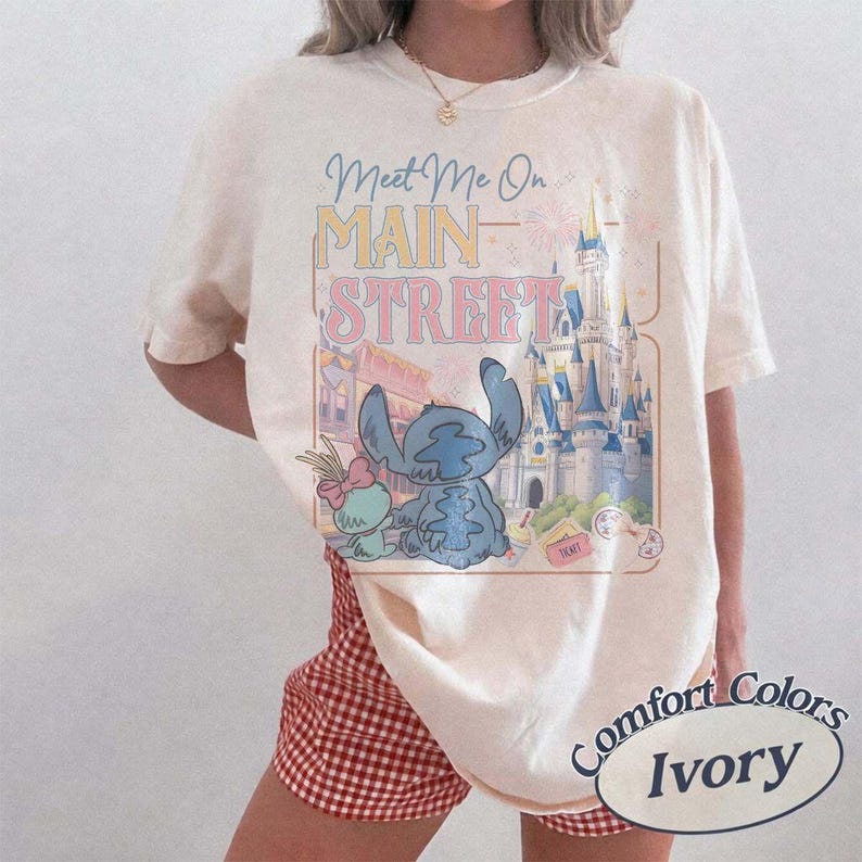May include: Ivory-colored t-shirt with a graphic featuring Stitch, a castle, and the text "Meet Me On Main Street." The design includes pastel colors and a vintage aesthetic. The shirt is a Comfort Colors brand.