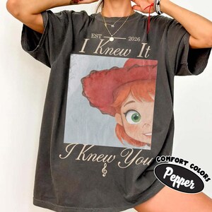 May include: A charcoal gray t-shirt featuring a graphic print of a character with red hair and a red hat. The text reads EST 2026 I Knew It and I Knew You with a small musical note symbol. The shirt has a relaxed fit and short sleeves.