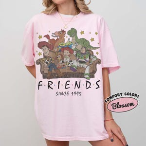 May include: A light pink t-shirt featuring a graphic print of Toy Story characters sitting on a couch with the text "FRIENDS SINCE 1995" below. The design includes Woody, Jessie, Buzz Lightyear, Rex, Bullseye, and the Aliens surrounded by gold stars.