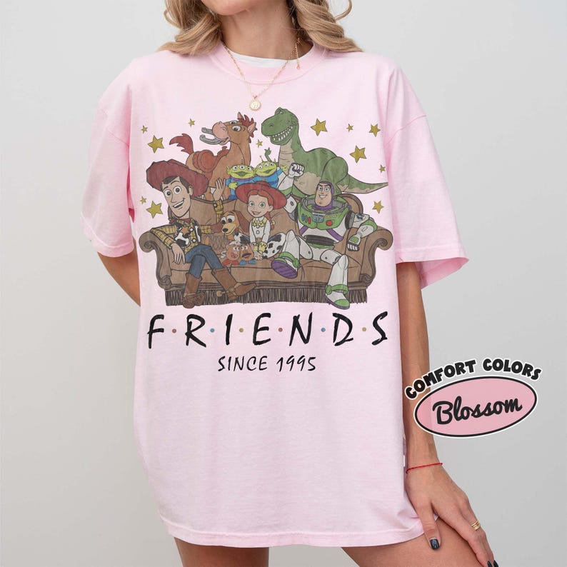 May include: A light pink t-shirt featuring a graphic print of Toy Story characters sitting on a couch with the text "FRIENDS SINCE 1995" below. The design includes Woody, Jessie, Buzz Lightyear, Rex, Bullseye, and the Aliens surrounded by gold stars.