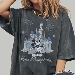 May include: Dark grey t-shirt with a graphic of Mickey Mouse and Cinderella's castle, fireworks, and the text "Walt Disney World." The shirt is a Comfort Colors Pepper tee.